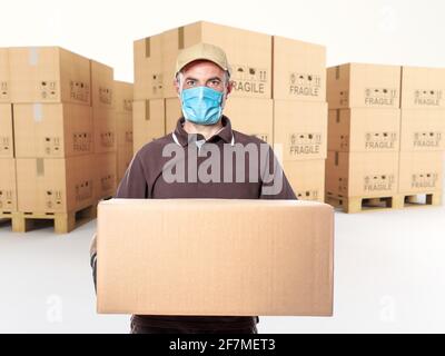 Kurier mit Maske und Paket in der Hand, Palette von Kartons im Hintergrund. Stockfoto