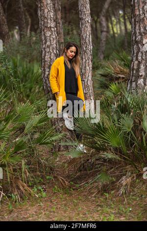 Frau, die im Frühjahr in einem Baum mit einem gelben Mantel im Wald oder Park sitzt. Hut auf dem Baum Stockfoto