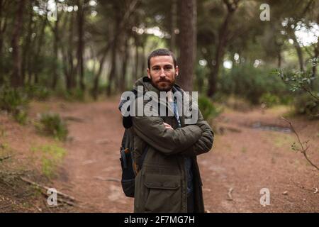 Mann im Wald mit gekreuzten Armen und einem grünen Mantel. Wandern. Er trägt einen Rucksack auf dem Rücken und eine Mütze. Stockfoto