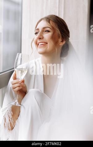 Braut s Morgen. Braut trinkt Champagner im Peignoir. Junge Frau sitzt auf einem großen Fenster in einem Hotelzimmer im Badezimmer. Schönes Mädchen in whi Stockfoto