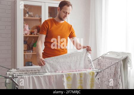 Mann, der Hausaufgaben macht. Der kaukasische Mann entfernt nach der Wäsche die Kleidung und die Babywäsche aus dem tragbaren Trockner im Wohnzimmer Stockfoto