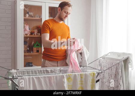 Mann, der Hausaufgaben macht. Der kaukasische Mann entfernt nach der Wäsche die Kleidung und die Babywäsche aus dem tragbaren Trockner im Wohnzimmer Stockfoto