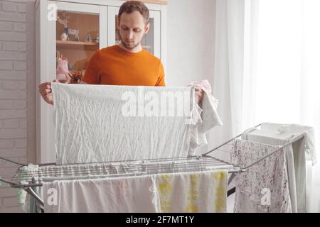 Mann, der Hausaufgaben macht. Der kaukasische Mann entfernt nach der Wäsche die Kleidung und die Babywäsche aus dem tragbaren Trockner im Wohnzimmer Stockfoto