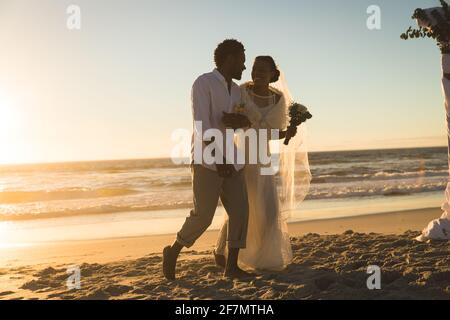 afroamerikanisches Paar, das verliebt heiratet und am Strand die Hände beim Sonnenuntergang hält Stockfoto