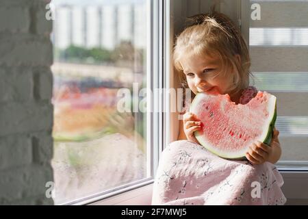 Kleine kaukasische Mädchen in einem rosa Kleid sitzt auf dem Fenster und frisst ein großes Stück Wassermelone Stockfoto