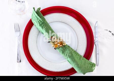 Eine grüne Serviette mit einem goldenen Serviettenring auf einer weißen Keramikplatte und weißem Tischtuch. Ort für ein Weihnachtsessen oder ein festliches Fest. Stockfoto