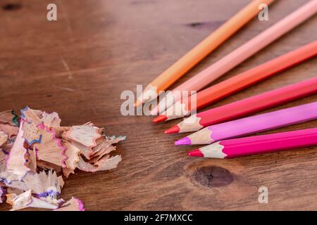 Buntstiftspangen, die aus einem blauen Bleistiftspitzer ohne Deckel auf einen Hartholztisch auslaufen, Farben sind violett, orange, rosa, gelb, Weichfokus. Stockfoto