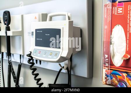 London, Ontario, Kanada - Februar 26 2021 - Nahaufnahme eines hängenden Blutdruckmonitors und einer Schachtel mit chirurgischen Handschuhen in einer kleinen Klinik. Stockfoto