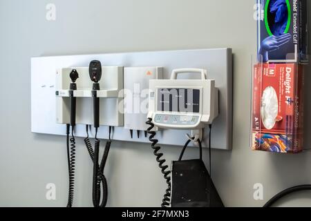 London, Ontario, Kanada - Februar 26 2021 - Nahaufnahme eines hängenden Blutdruckmonitors und einer Schachtel mit chirurgischen Handschuhen in einer kleinen Klinik. Stockfoto