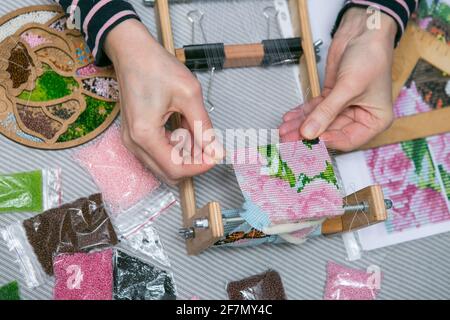 Weben aus Perlen. Nahaufnahme - die Hände einer Frau ziehen Perlen an einen Faden und machen Schmuck. Die Frau webt gern aus Perlen. Stockfoto