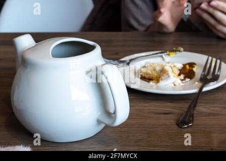 Ein weißer Keramikbecher neben einem Teller Kuchenbrösel mit zwei silbernen Gabeln auf einem Hartholztisch, Februar 2021, in einem kleinen lokalen Café. Stockfoto