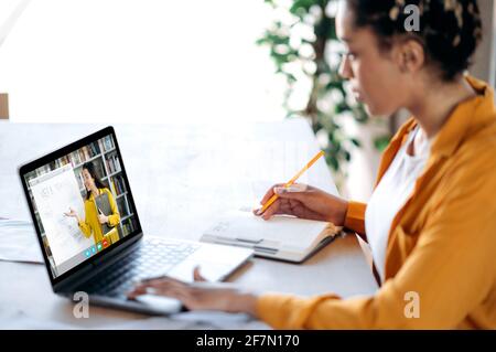 Fernunterricht, Online-Vorlesung. Afroamerikanische Studentin, die zu Hause mit einem Laptop studiert, sich eine Online-Lektion anhört, zeigt die Lehrerin auf dem Laptop-Bildschirm Informationen auf einem Whiteboard Stockfoto