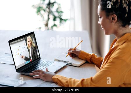 Fernunterricht per Videokonferenz. Afroamerikanische Studentin, die zu Hause mit einem Laptop lernt, hört sich eine Online-Lektion an, auf dem Laptop-Bildschirm zeigt die Lehrerin Informationen auf dem Whiteboard Stockfoto