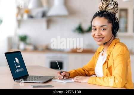 E-Learning-Konzept. Glückliche afroamerikanische Schülerin mit Dreadlocks, die ein Headset trägt, sitzt zu Hause am Schreibtisch, macht sich Notizen, studiert aus der Ferne, schaut freundlich lächelnd auf die Kamera. Online Stockfoto