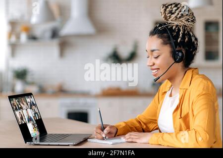 Eine glückliche afroamerikanische Studentin hört sich einen Online-Vortrag mit einem Laptop und einem Headset an, nimmt Notizen, studiert aus der Ferne, die Lehrerin und andere Schüler sind auf dem Laptop-Bildschirm. E-Learning Stockfoto