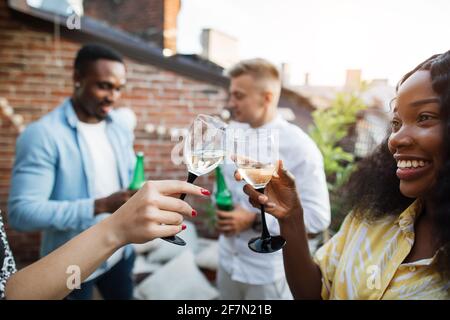 Zwei Frauen toasten mit einem Glas Champagner, während zwei Männer Bier trinken und sich im Hintergrund unterhalten. Vier gemischte Rennfreunde genießen Party-Zeit auf dem Dach. Stockfoto