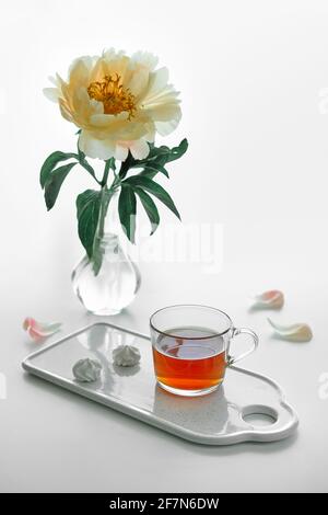 Tasse Tee und weiße Salmler auf Keramik-Tablett. Einzelne Pfingstrose Blume in trendigen modernen Glasvase auf weißem Tisch. Blassgelbe, cremefarbene Blütenblätter und Stockfoto