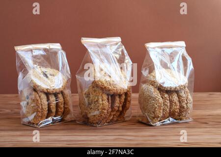 Drei Arten von Haferflocken - mit Schokolade, Walnüssen und Rosinen und Sesamsamen - in transparenten Tüten auf einem Holztisch an einer braunen Wand. C Stockfoto