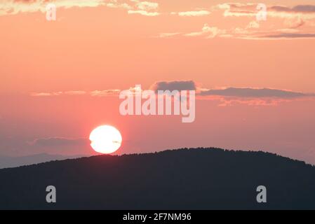 Erstaunlicher zartrosa Sonnenuntergang mit einer runden Sonnenscheibe über Die Konturen von Bergen bedeckt mit Wald und beleuchteten Wolken Am Himmel Stockfoto