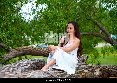 Ganzkörperexposition Erwachsene philippinische Frau in Kleid Blick auf die Kamera Während Sie sich am Sommertag auf dem Baumstamm entspannen Wald Stockfoto