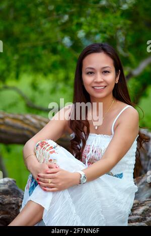 Ganzkörperexposition Erwachsene philippinische Frau in Kleid Blick auf die Kamera Während Sie sich am Sommertag auf dem Baumstamm entspannen Wald Stockfoto