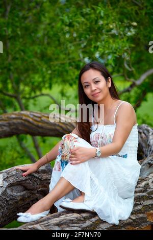 Ganzkörperexposition Erwachsene philippinische Frau in Kleid Blick auf die Kamera Während Sie sich am Sommertag auf dem Baumstamm entspannen Wald Stockfoto