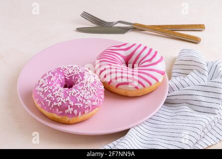 Zwei zuckerbeschichtete Donuts liegen auf einer rosa Keramikplatte. Servieren mit Besteck und Leinenservietten. Selektiver Fokus. Stockfoto