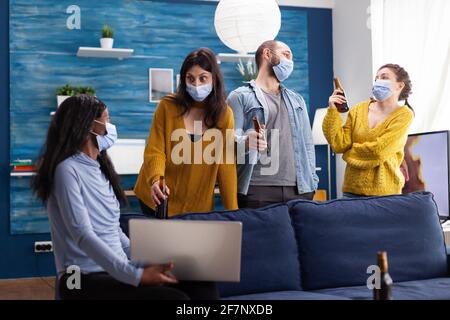 Multirassische Gruppe von Freunden, die mit einem Laptop chatten und dabei soziale Distanz bewahren und eine Gesichtsmaske tragen, um die Ausbreitung von Covid und Infektionen im Wohnzimmer der Wohnung zu verhindern. Konzeptuelles Image Stockfoto