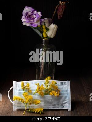Eine Schutzmaske gegen Covid-19 ist mit gelben Blüten von Sedum palmeri verziert, mit einer Glasvase mit getrockneten Blumen im Hintergrund Stockfoto