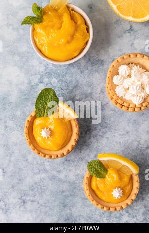Minitörtchen mit Zitronenquark, Mini-Baiser, Zitronenscheiben und Minze auf blauem Betongrund. Draufsicht. Selektiver Fokus. Stockfoto