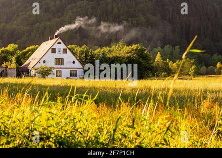 Ländliche Aussicht, ein traditionelles polnisches Landhaus mit Rauch aus dem Kamin neben Kiefernwald. Landhaus in der Ferne bei Sonnenuntergang Stockfoto