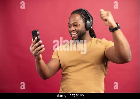 Froher, dunkelhäutiger, prall gelaunter Mann tanzt, macht Bewegungen zur Musik, hat moderne Stereokopfhörer, lächelt positiv, ist in Hochstimmung, hält Mobiltelefon in der Hand. Sorgloser Kerl hört lebendige Musik Stockfoto