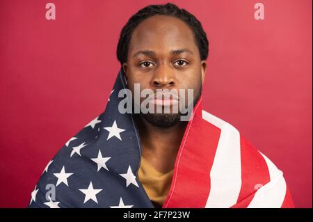 Close Portrait of serious trauriges hurted dark skinned american bärded man covered with usa flag, Head shot, looking at camera, isolated over Red background, depends his equal rights, lives in Freedom Stockfoto