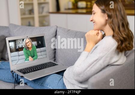 Seitenansicht eine frohe junge Frau, die einen Laptop-Computer zur Videoverbindung mit einer älteren Frau in der Ferne benutzt. Erwachsene Tochter ruft per Video an ältere grauhaarige Mutter auf dem Bildschirm Stockfoto