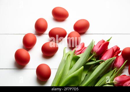 Ostereier und rote Blumen auf weißem Holztisch. Bemalte Hühnereier und rote Tulpen, Einrichtung am Ostertag, selektiver Fokus Stockfoto