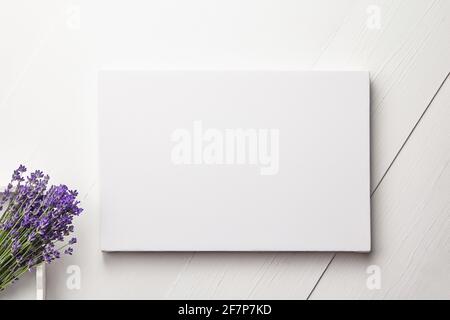 Weißes Canvas-Mockup und Lavendelblüten-Bouquet auf weißem Holztisch-Hintergrund, flach liegend. Leere Leinwand, Draufsicht Stockfoto
