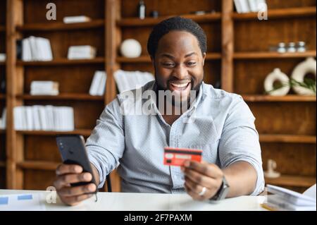 Überglücklich afroamerikanischer Unternehmer in einem eleganten Casual-Hemd hält Telefon und Bankkarte, macht eine Online-Bestellung, bezahlt für die Lieferung von Lebensmitteln im Büro, online einkaufen, E-Banking-Konzept Stockfoto