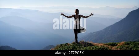 Silhouette einer jungen Frau, die Yoga-Pose auf einem grasbewachsenen Hügel durchführt und wunderschöne Berge anschaut. Sportliche Frau, die morgens auf einem Bein steht, während sie im Freien Yoga praktiziert. Stockfoto