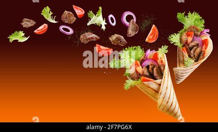 Zwei Döner-Kebab oder Shawarma mit Zutaten, die in der Luft schweben: Rindfleisch, Salat, Zwiebeln, Tomaten, Gewürz. Orangebrauner Hintergrund. Speicherplatz kopieren. Stockfoto