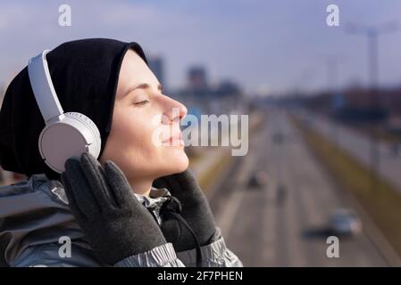 Lächelnd schöne junge Erwachsene Frau hören Musik mit ihr Smartphone und Kopfhörer zu Fuß durch die Stadt Stockfoto