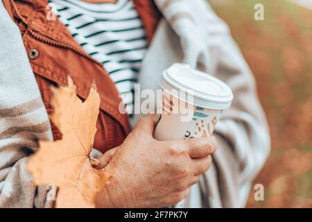 Kaukasische Frau hält Hand mit Herbst Ahornblatt und Papierbecher mit heißem Getränk. Dame in warmer Decke in der Herbstsaison Stockfoto