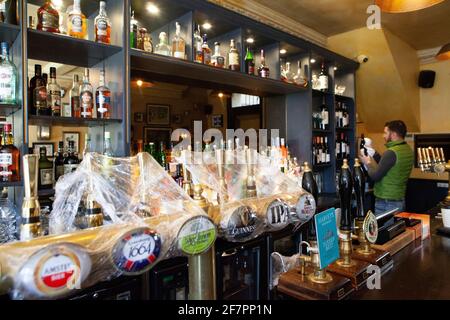 London, Großbritannien, 9. April 2021: Der Abbeville Pub in Clapham wird ab dem 12. April wieder für Drinks im Freien und zum Abendessen auf der Terrasse geöffnet. Während die Bierzapfhähne unter Plastikfolie sauber gehalten werden, stäubt Barkeeper Klodian Flaschen mit Spirituosen. Anna Watson/Alamy Live News Stockfoto