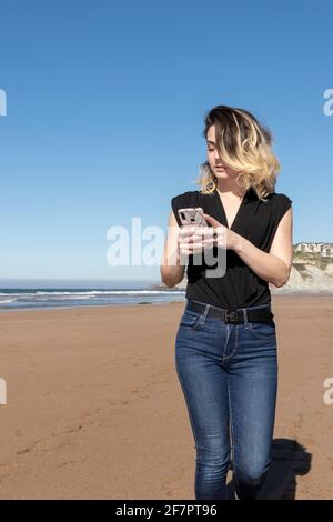 Junge blonde Frau, die am Strand läuft und die anschaut Telefonieren Sie an einem sonnigen Frühlingstag mit blauem Himmel Stockfoto
