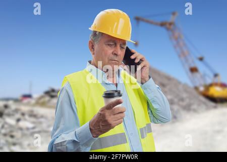 Alter Konstrukteur, der bei der Arbeit auf dem Smartphone spricht hardhat und Weste halten Kaffee in Take-away Papierbecher mit Baustelle Hintergrund Stockfoto