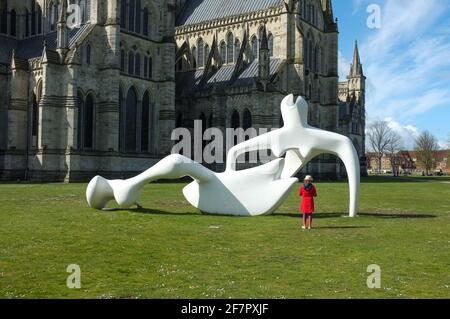 Große Reclining Figure Skulptur von Henry Moore, die von einem jungen Mädchen in Rot in der Salisbury Cathedral Close in Wiltshire untersucht wurde. VEREINIGTES KÖNIGREICH. April 2021. Stockfoto