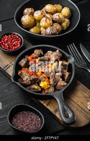 Rindfleisch, geschmort mit Kartoffeln, Karotten und Gewürzen, in gusseiserner Pfanne, auf schwarzem Holzhintergrund Stockfoto