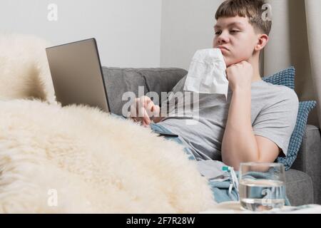 Teenager sitzt auf der Couch und spielt beim Online-Lernen auf dem Laptop während Quarantäne und Krankheit. Selbstisolierung. Quarantäne. E-learning über Entfernungen Stockfoto