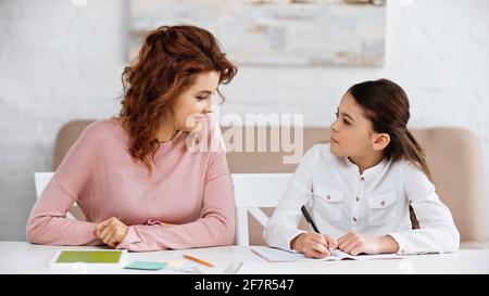 Lächelnde Frau, die in der Nähe des Kindes sitzt und auf einem Notebook und digital schreibt Tablet mit grünem Bildschirm Stockfoto