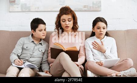 Kinder mit digitalem Tablet und Notebook sitzen in der Nähe der Mutter beim Lesen Zu Hause buchen Stockfoto