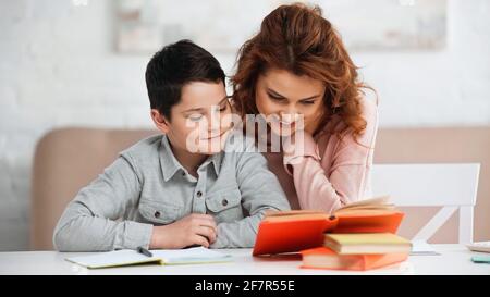 Lächelnde Frau, die in der Nähe des präteen Sohnes sitzt und Bücher über verschwommen Vordergrund Stockfoto